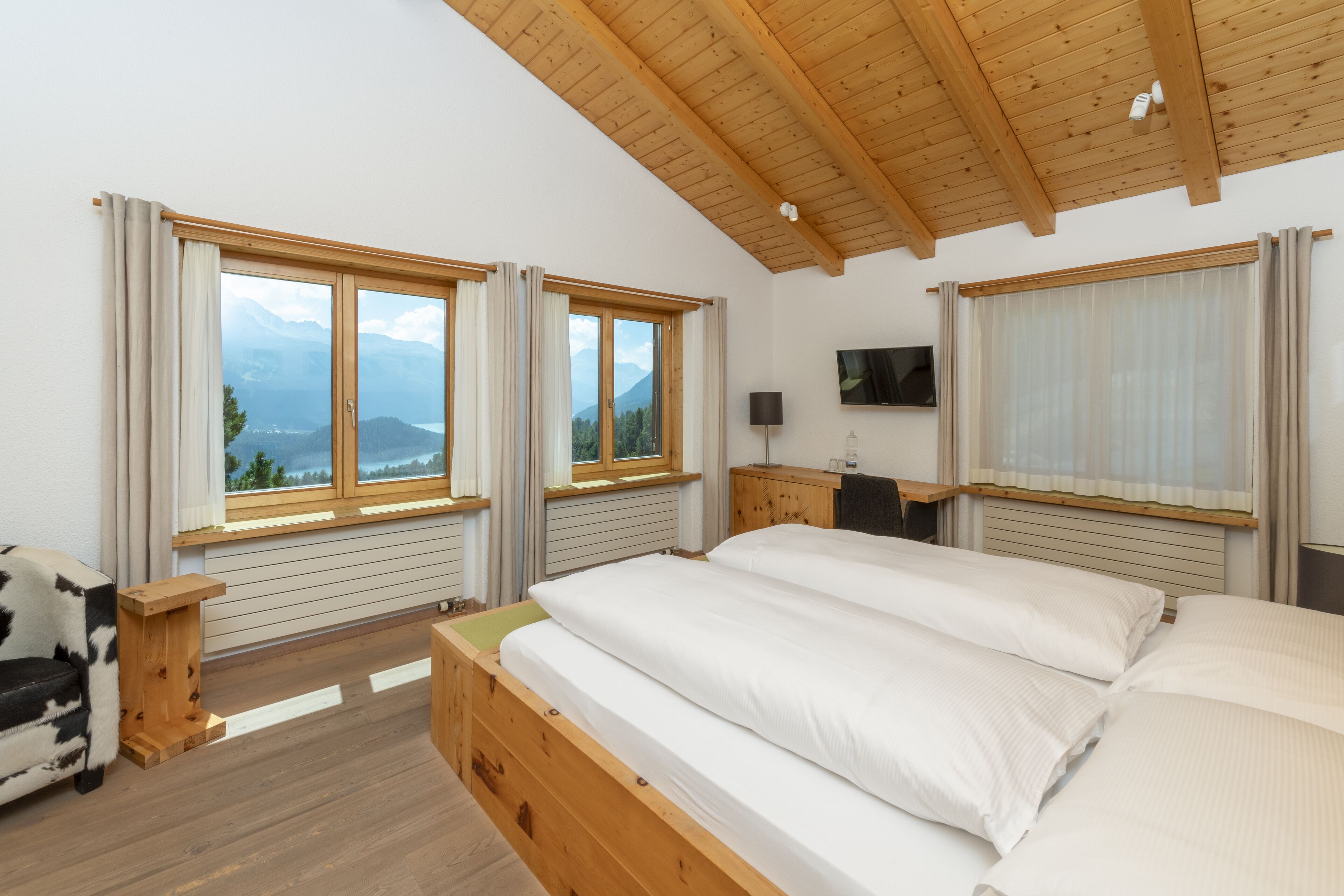 A bedroom with a wooden ceiling, two windows with curtains, a bed, a desk, and a TV.