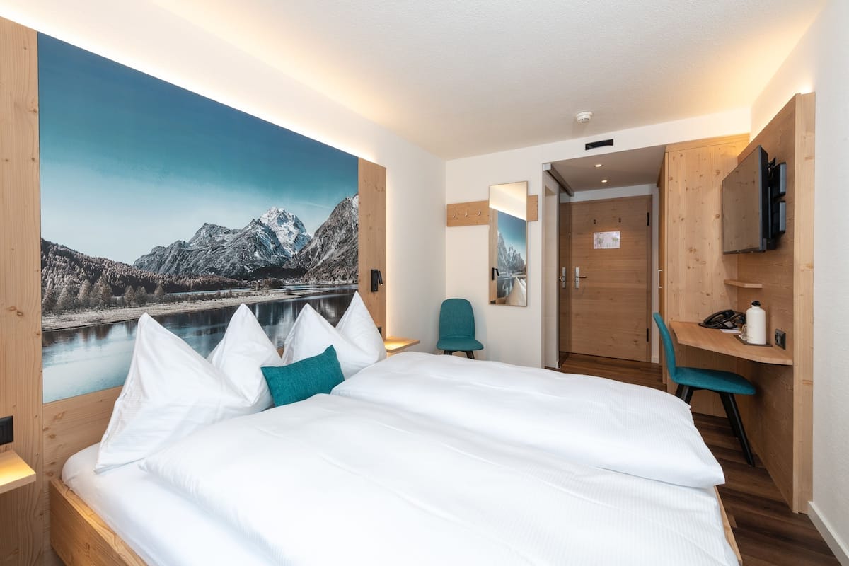 A hotel room with a large mountain view mural, a bed with white linens, a teal chair, and a wooden desk with a flat-screen TV.
