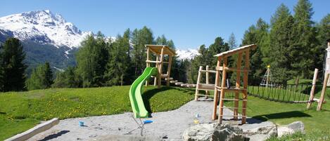 Un parc infantil amb tobogan i elements per escalar, amb les muntanyes nevades i els pins al fons.