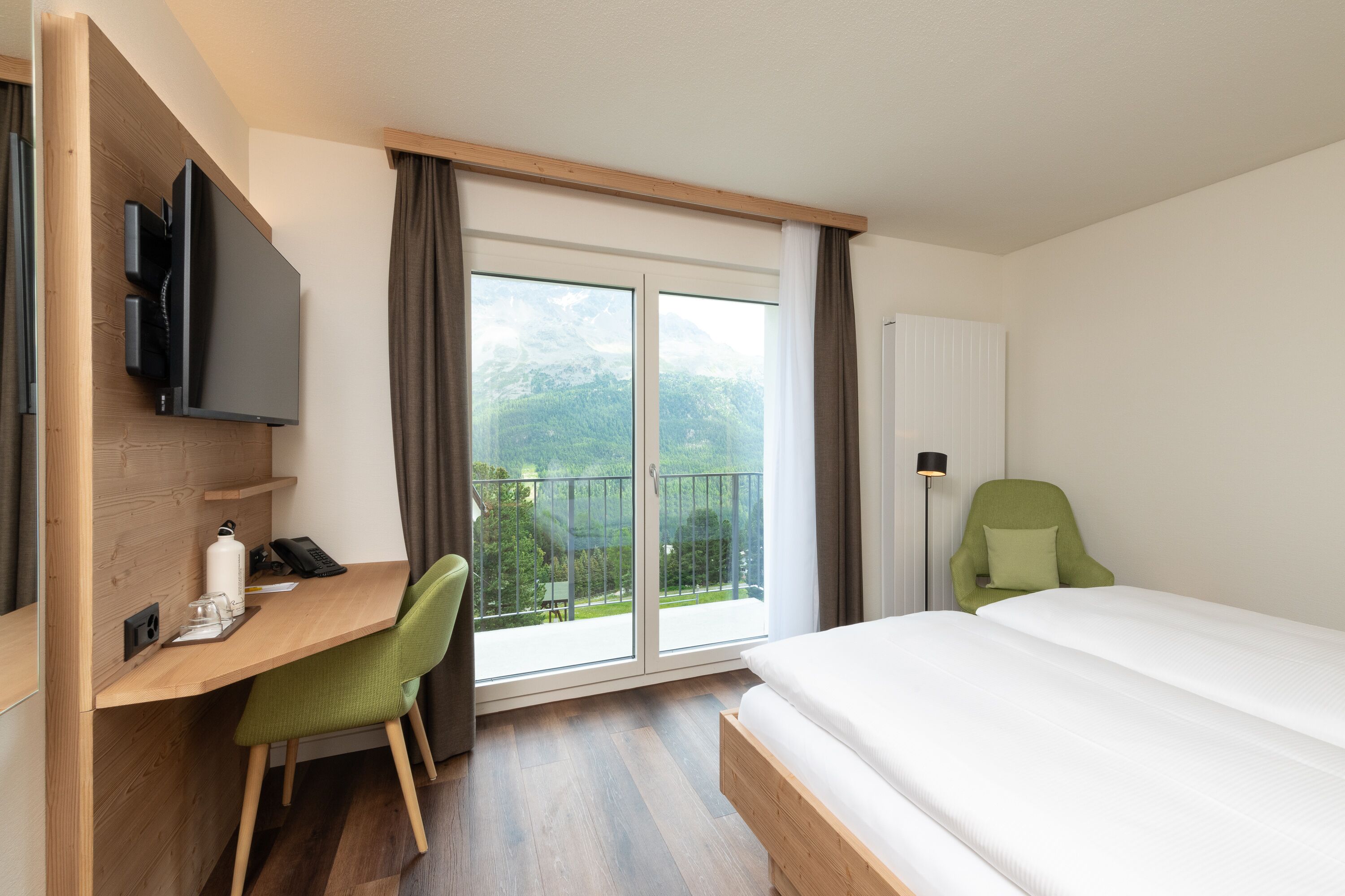 A hotel room with a bed, a desk with a chair, a television, and a view of mountains through a large window.