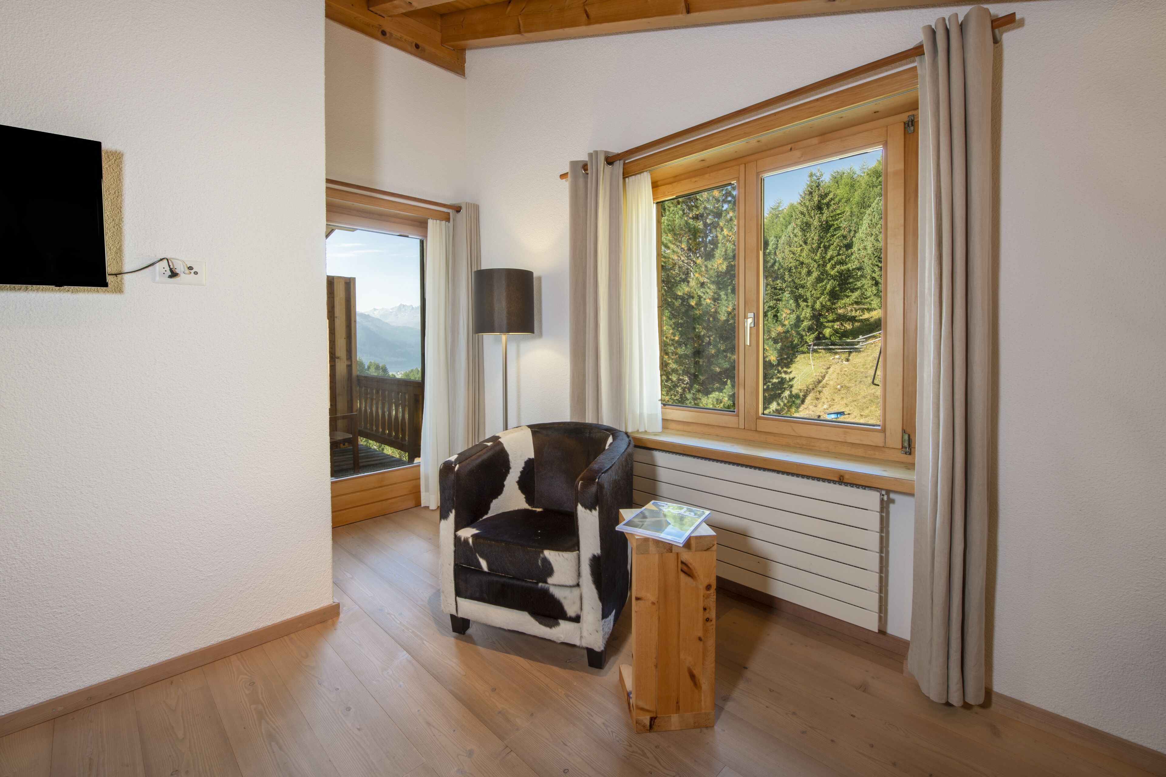 Una habitación acogedora con suelo de madera, un sillón de estilo clásico en blanco y negro, una pequeña mesita de madera, una lámpara y un amplio ventanal con cortinas.