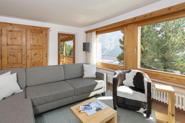 A modern living room with a grey sofa, wooden cabinets, a wooden chair, and a large window offering a mountain view. - Berghotel Randolins (St. Moritz)