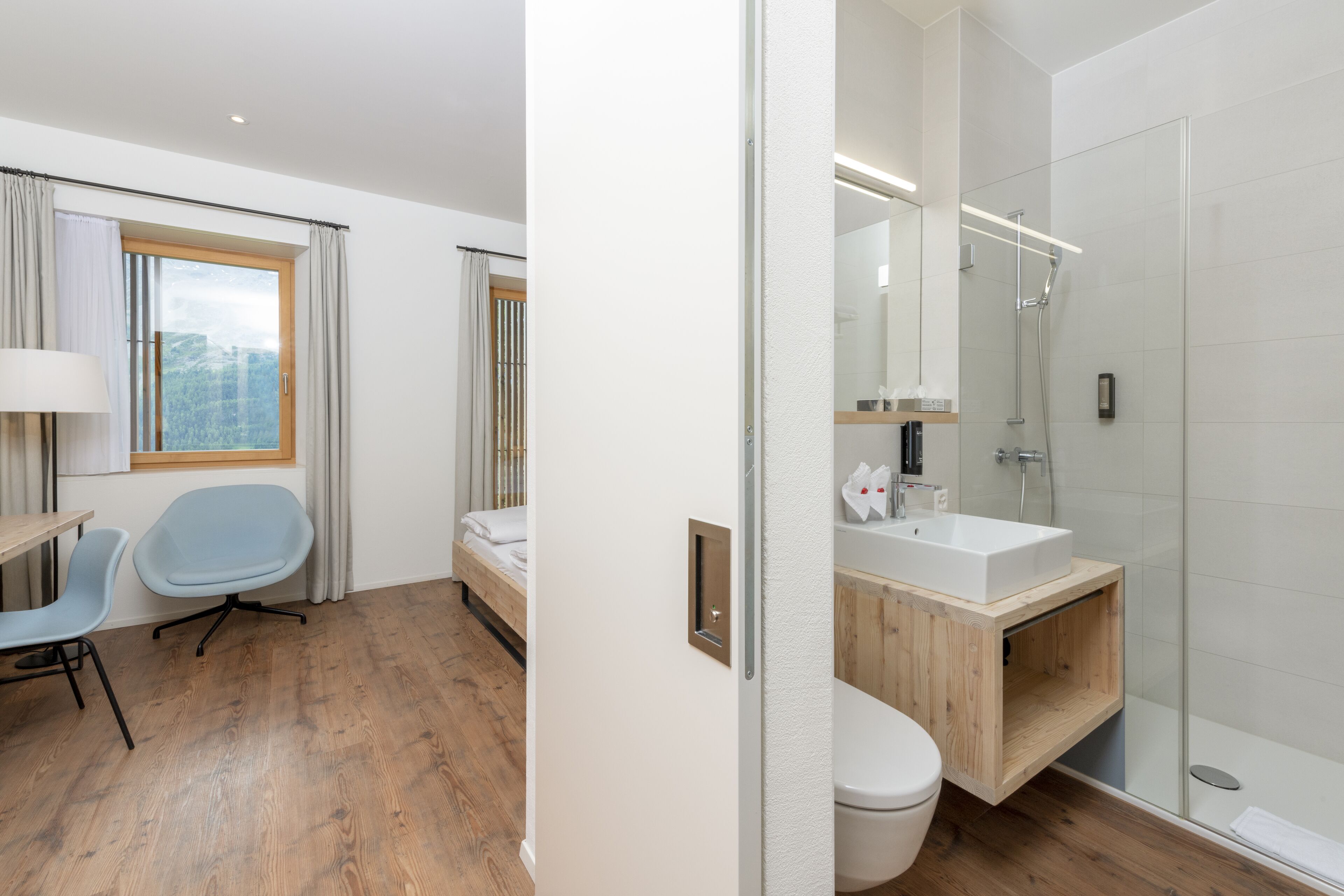 A modern hotel room with a wooden floor, a bed, a desk with a chair, a bathroom with a shower, and a large window with curtains.