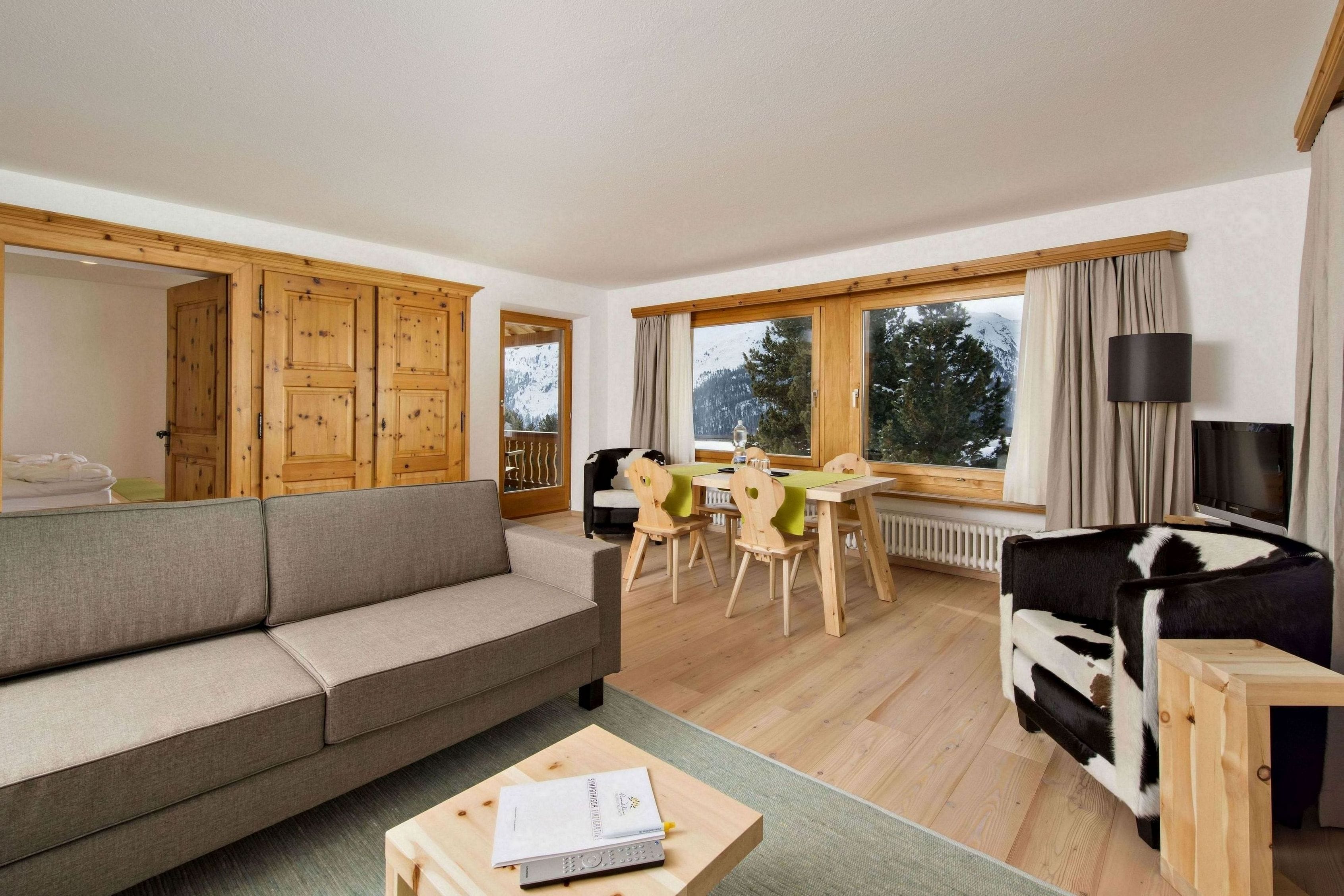 A cozy living room with a sofa, a wooden coffee table, a dining area with chairs, and a view of the outdoors through large windows.
