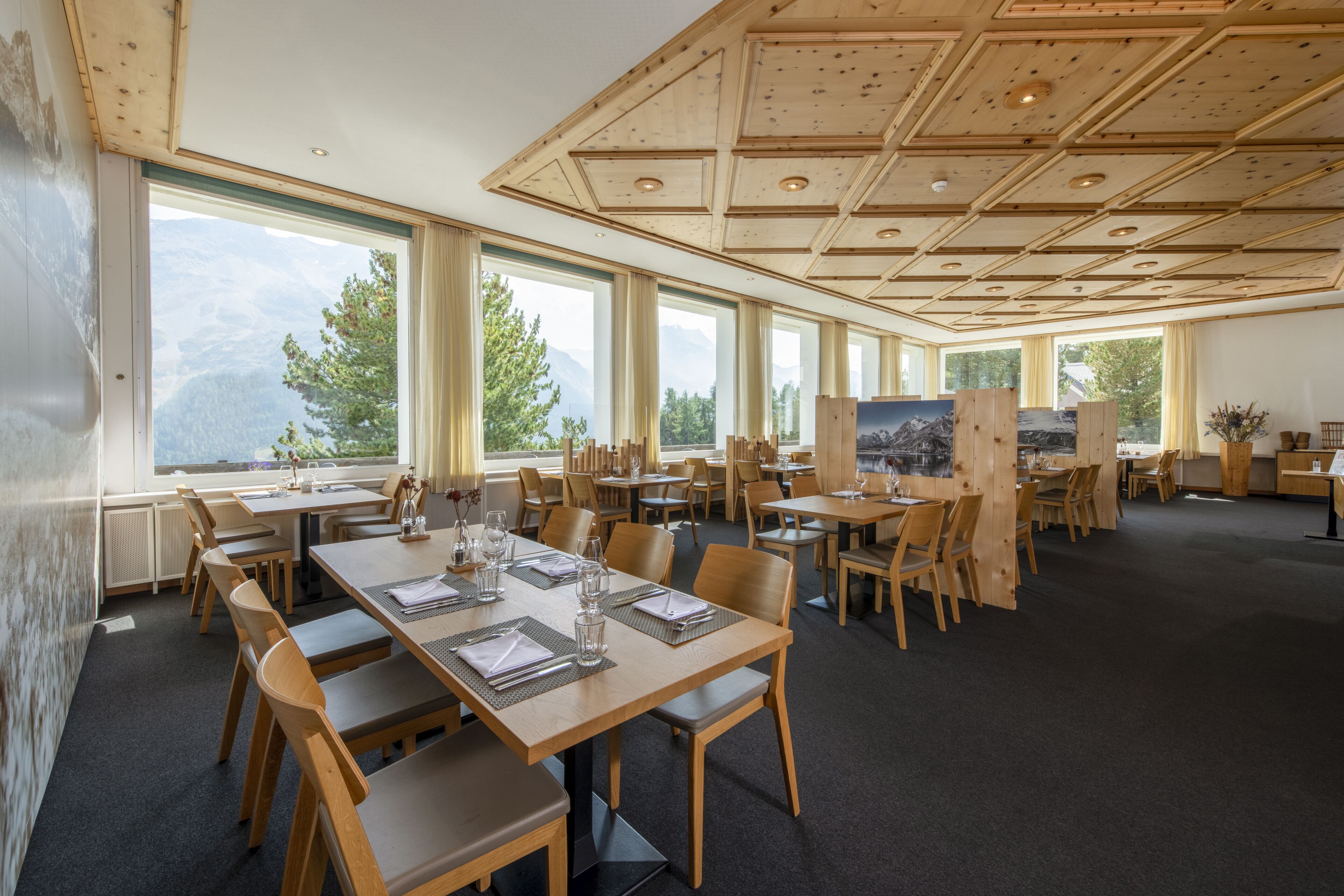 A spacious dining area with multiple wooden tables and chairs, large windows offering mountain views, and a high ceiling with wooden beams.