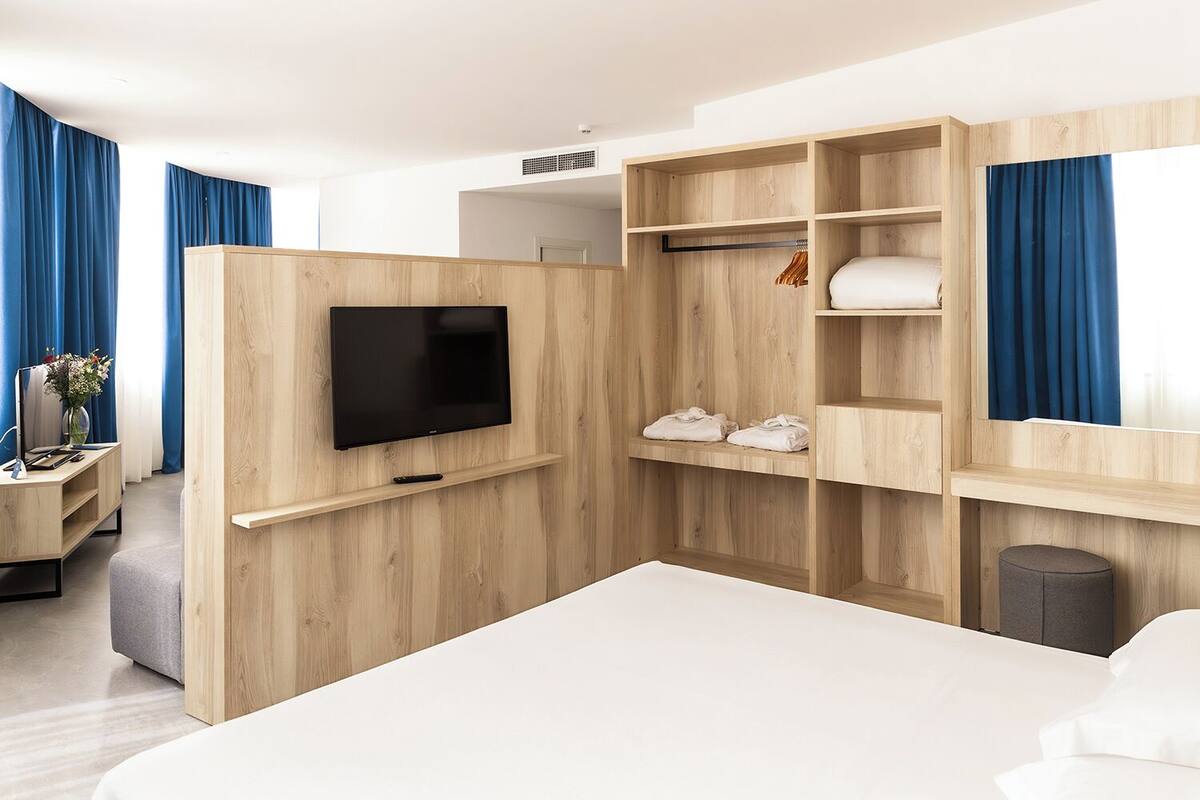 Une chambre d’hôtel moderne dotée d’un grand lit, d’un téléviseur fixé au mur en bois, d’un petit bureau avec une lampe et d’une armoire avec des étagères intégrées.
