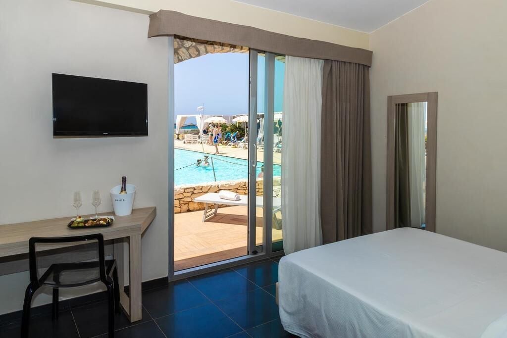 Hotelkamer met een bed, een televisie, een bureau met een stoel, een fles champagne en uitzicht op een zwembad en strand.