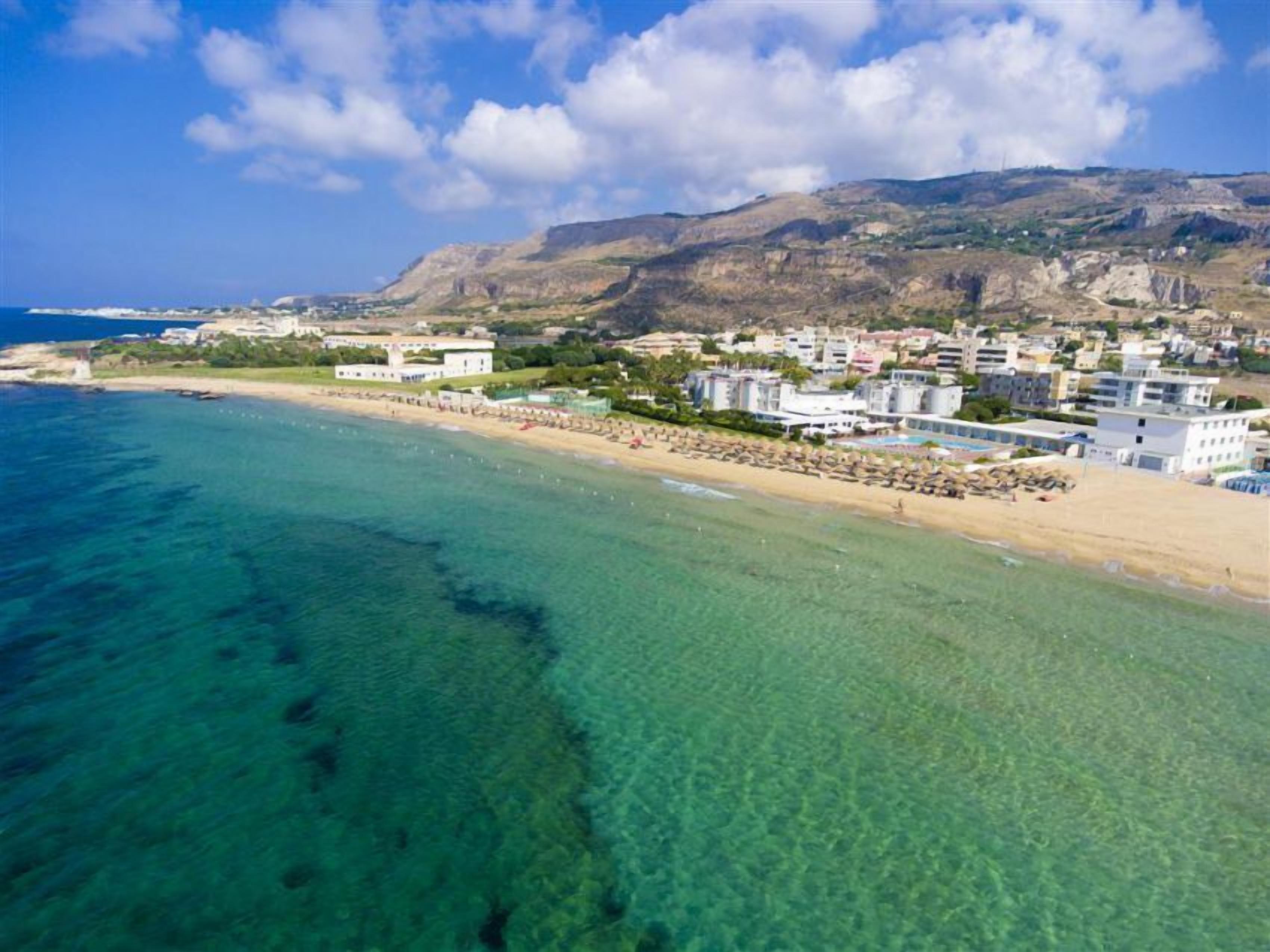 Vista aerea di una zona costiera con acque turchesi cristalline, spiaggia sabbiosa e un mix di edifici residenziali e vegetazione.