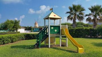 Un terrain de jeux avec un toboggan et une zone d’escalade, entouré de pelouse et de palmiers.