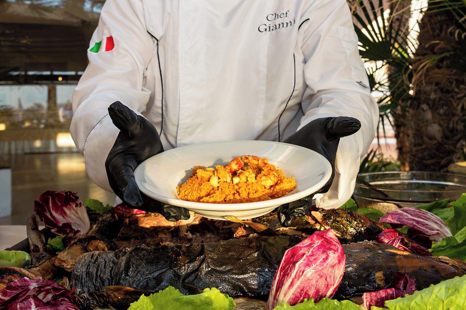 A chef in a white uniform with black gloves holding a plate of food.