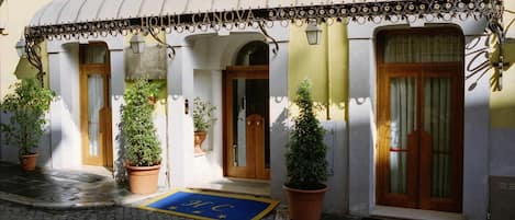 Entrada de hotel con porche cubierto, plantas en macetas y felpudo azul con un logotipo.
