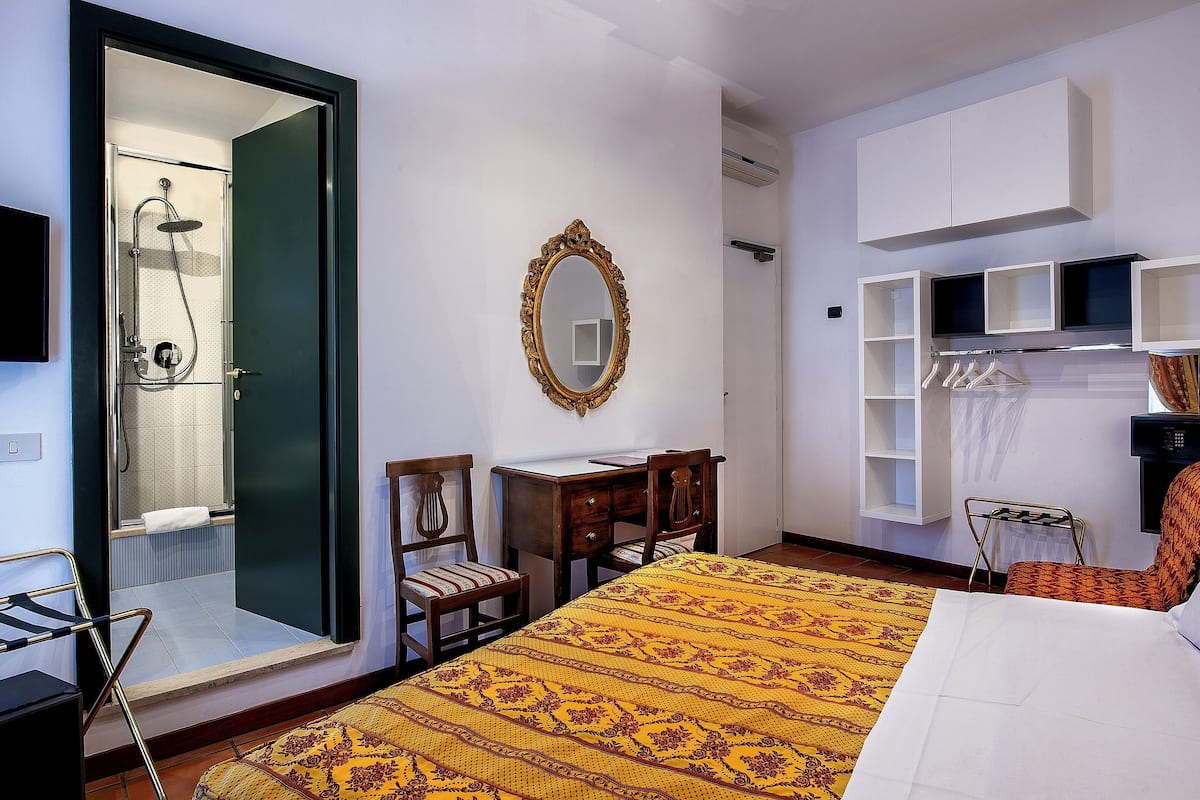 Habitación de hotel con cama, escritorio, silla, espejo, armario y zona de ducha.