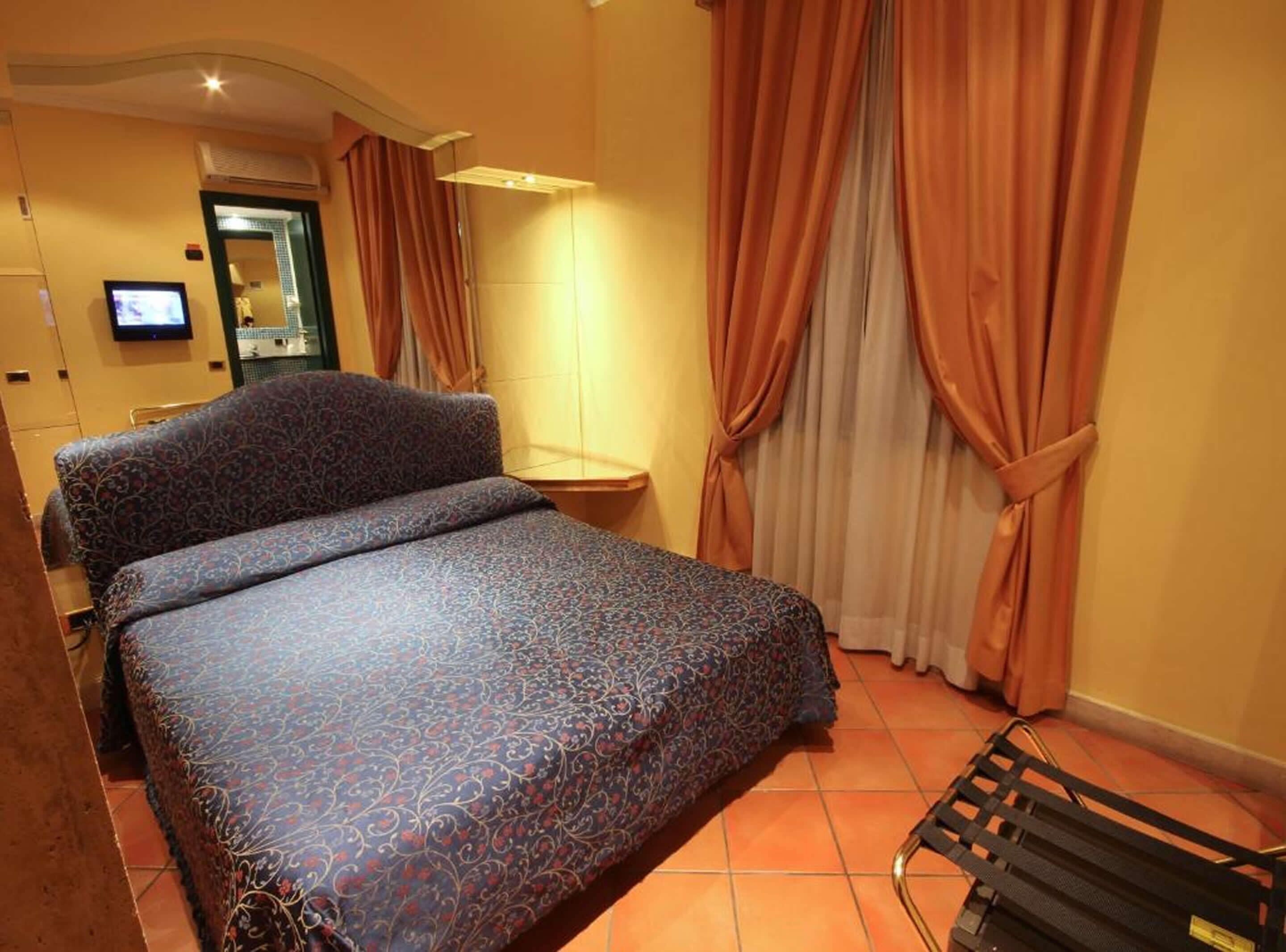 A hotel room with a bed, curtains, a TV, and a bathroom visible in the background.