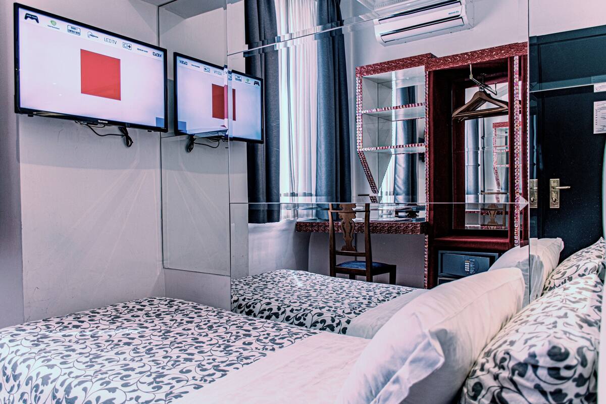 Habitación de hotel con dos camas, un escritorio, un espejo y dos monitores mostrando cuadrados rojos.