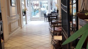 Een hotelgang met een betegelde vloer, een eetgedeelte met tafels en stoelen, en een uitzicht op de straat door grote ramen.