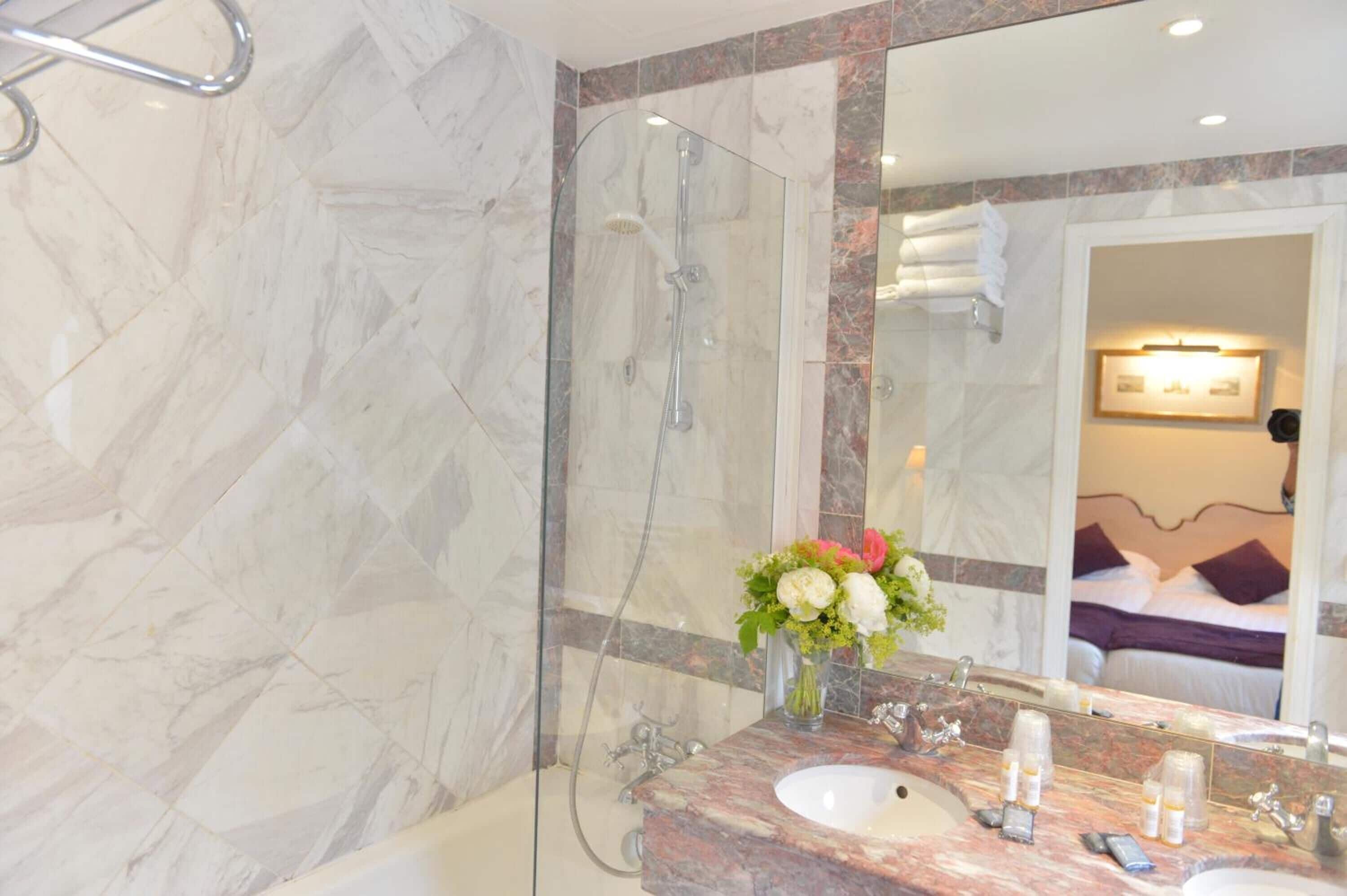 Une salle de bain avec un mur en marbre, une douche en verre, un lavabo agrémenté d’un vase de fleurs et un lit visible à travers un miroir.