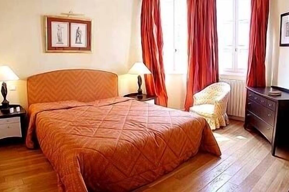 Ein Schlafzimmer mit einem großen Bett, zwei Nachttischen mit Lampen, einem Sessel, einem Schminktisch und zwei gerahmten Bildern an der Wand.