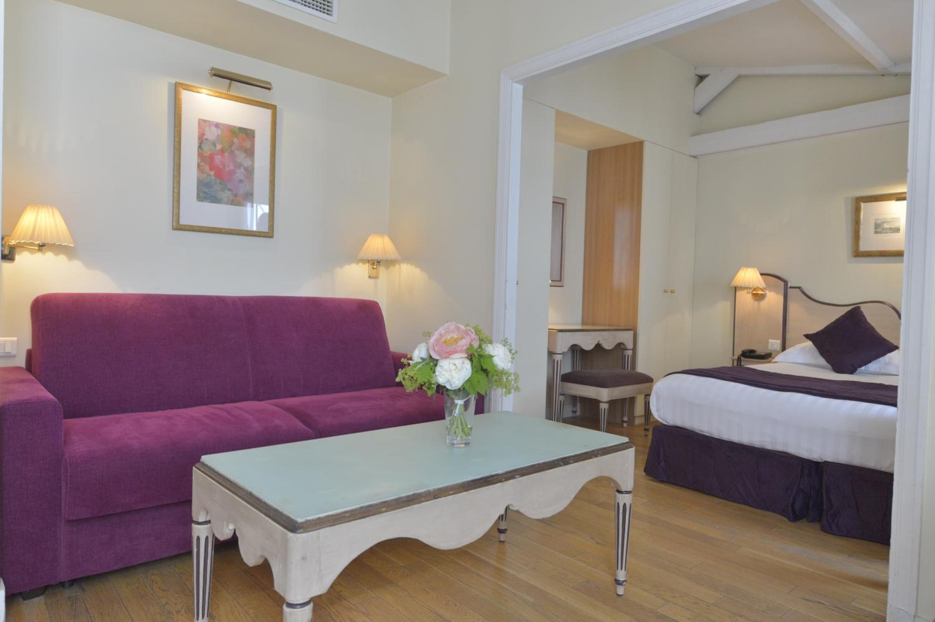 Ein Zimmer mit einem violetten Sofa, einem kleinen Tisch mit Blumenvase und einem Bett mit weiß-violetter Bettdecke, sowie einem Sessel.