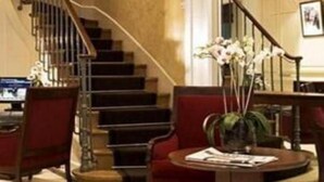 Een hotel lobby met een trap, rode stoelen, een ronde tafel met een bloemenvaas en een ingelijst schilderij aan de muur.