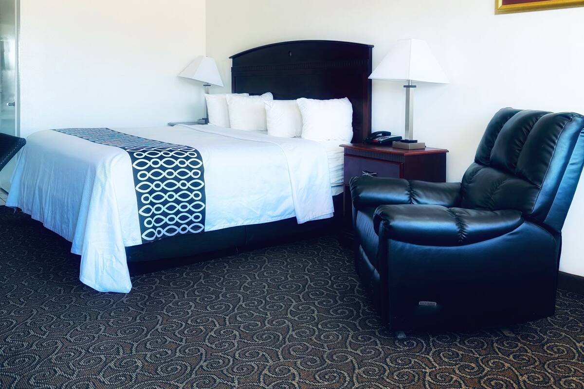 A hotel room with a bed, two bedside lamps, a black leather chair, and a patterned carpet.