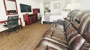 Habitación de hotel con un sofá de cuero marrón, un escritorio con una silla, un aparador, una cama con colcha estampada y un televisor en la pared.