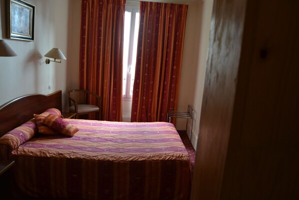 A bedroom with a bed, a chair, a lamp, and a window with curtains. - Hôtel Montpellier (Paris)