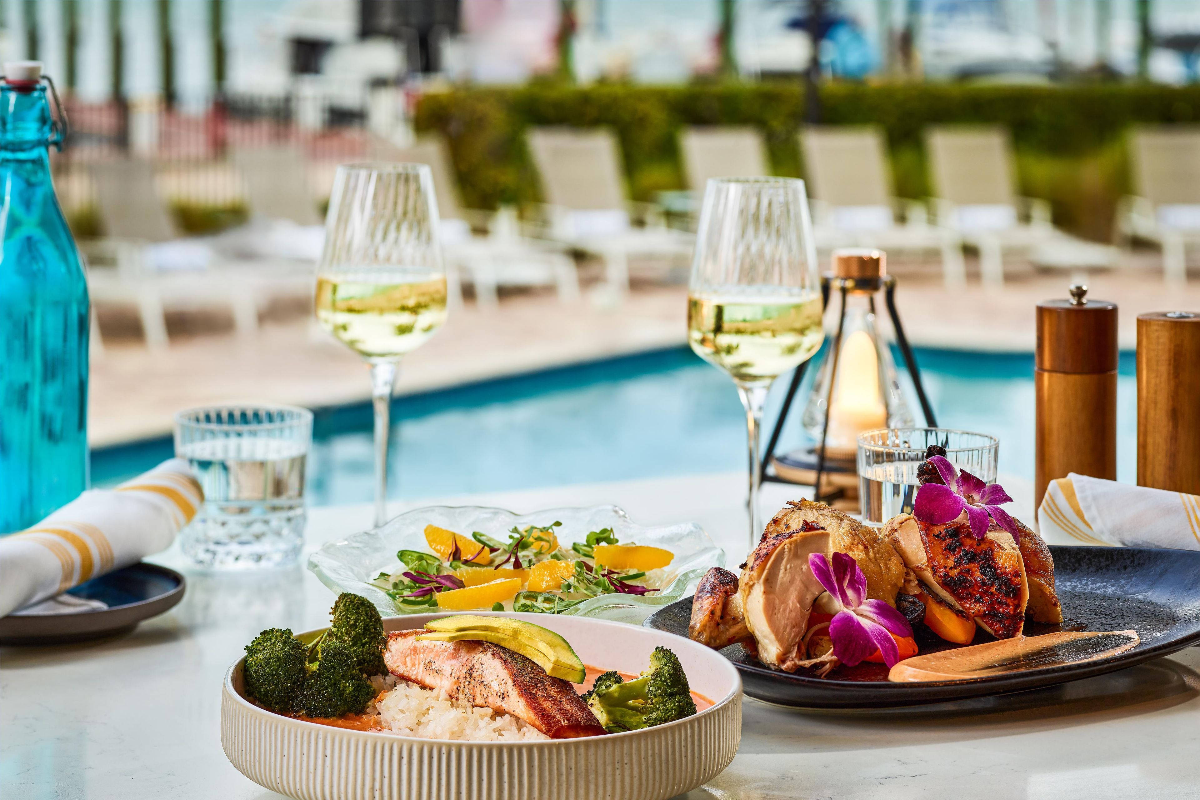 A dining table with a variety of dishes, including salmon and vegetables, near a poolside setting.
