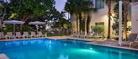 Una piscina con sillones de descanso y sombrillas, rodeada de palmeras y un edificio moderno.