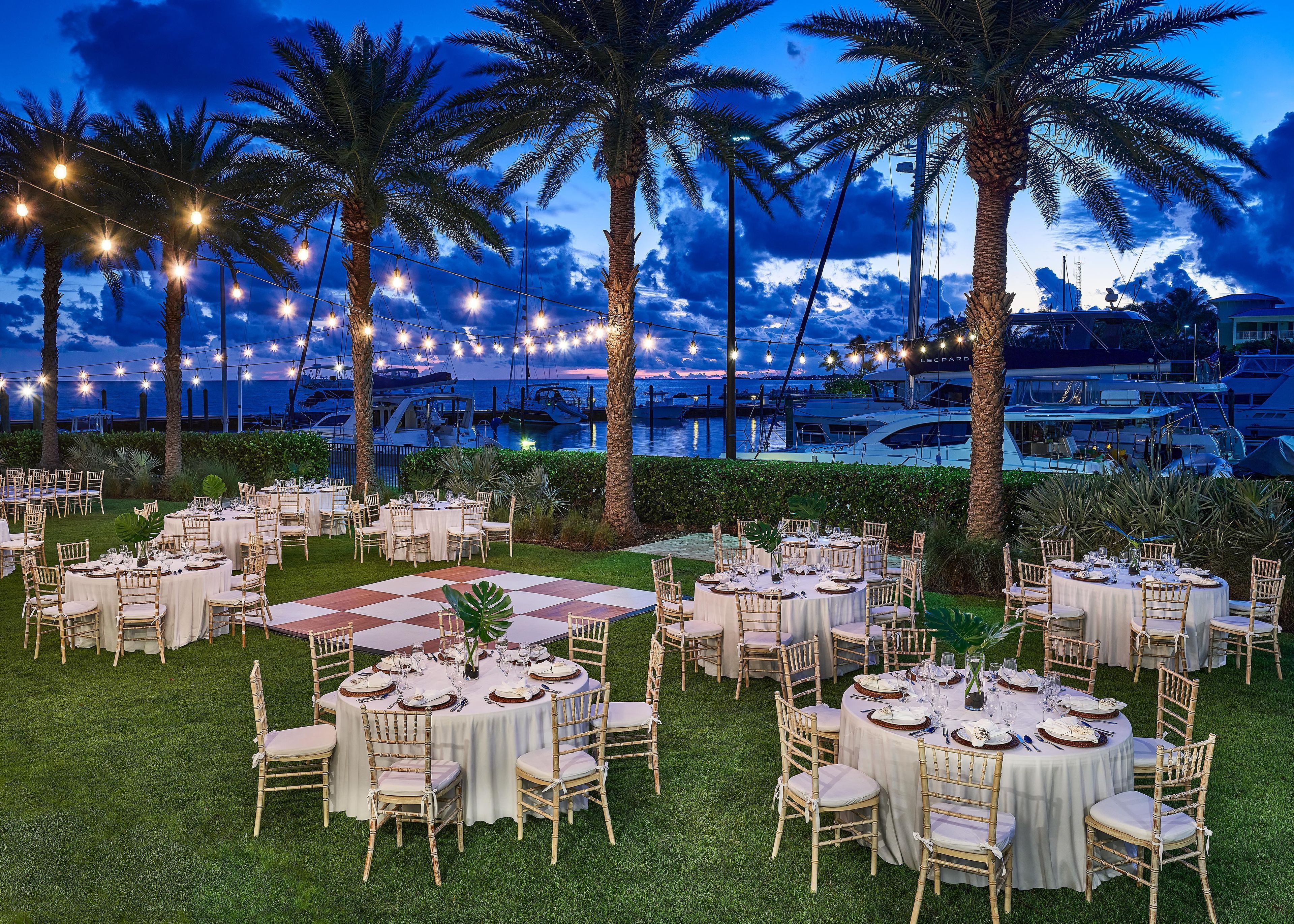 An outdoor dining area with round tables and chairs, string lights, and palm trees.