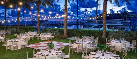 An outdoor dining area with round tables and chairs, string lights, and palm trees.