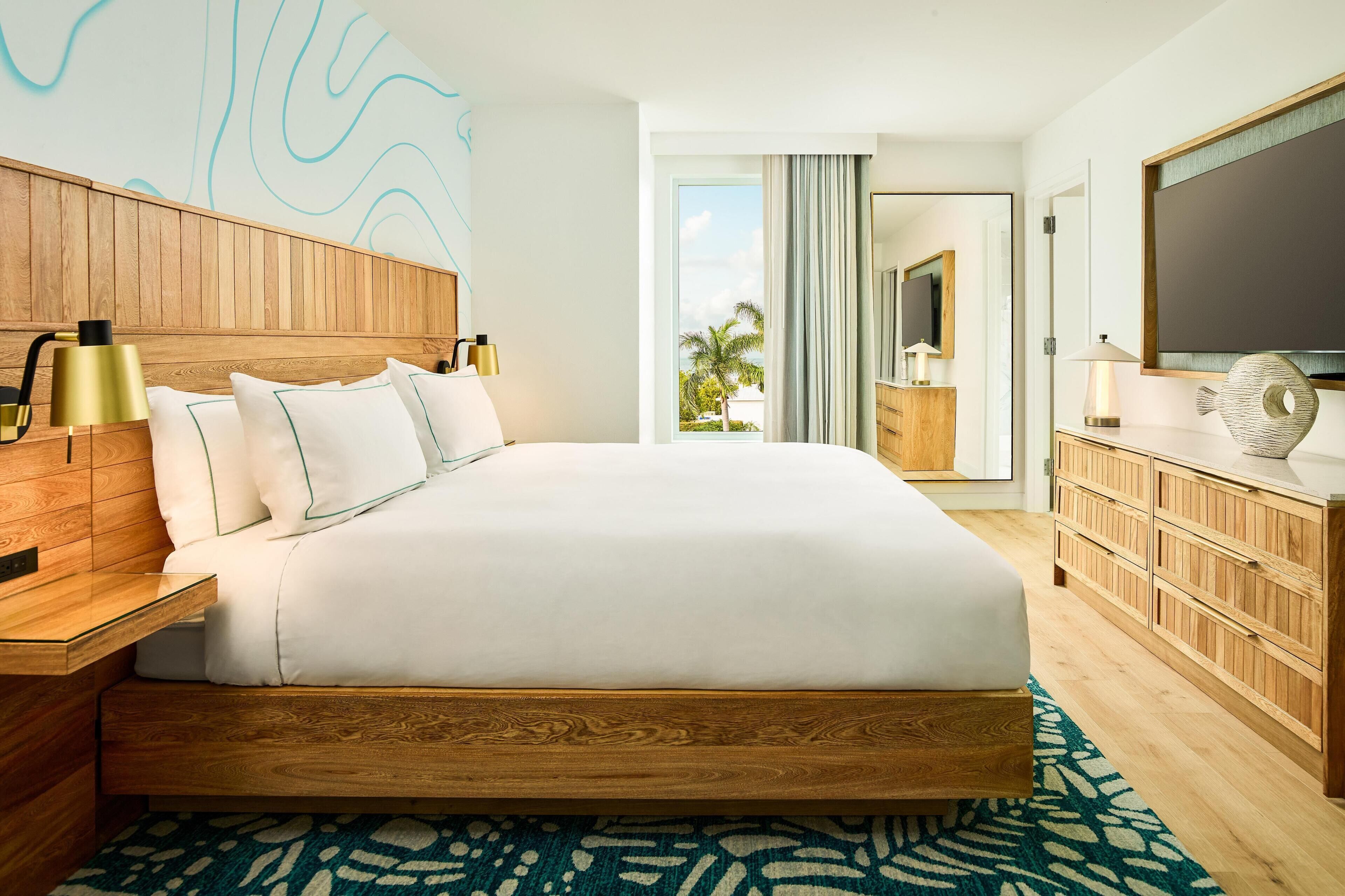 Une chambre d’hôtel avec un grand lit, une tête de lit en bois, une table de chevet, une commode, un téléviseur à écran plat et une vue sur les palmiers par la fenêtre.