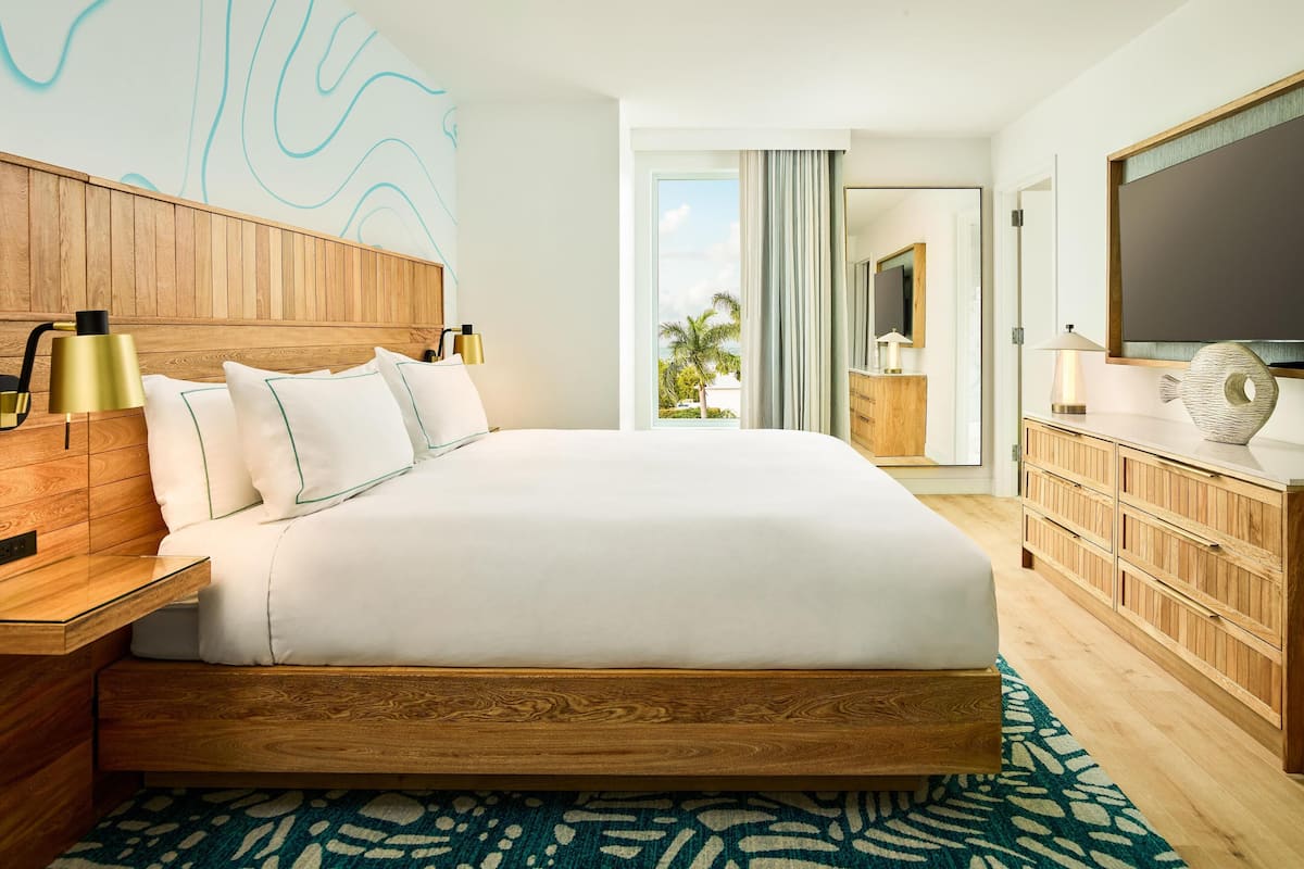 A hotel room with a large bed, wooden headboard, a nightstand, a dresser, a flat-screen TV, and a view of palm trees through the window.