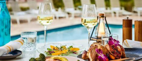 Mesa de comedor con comida, copas de vino blanco y ambiente junto a la piscina.