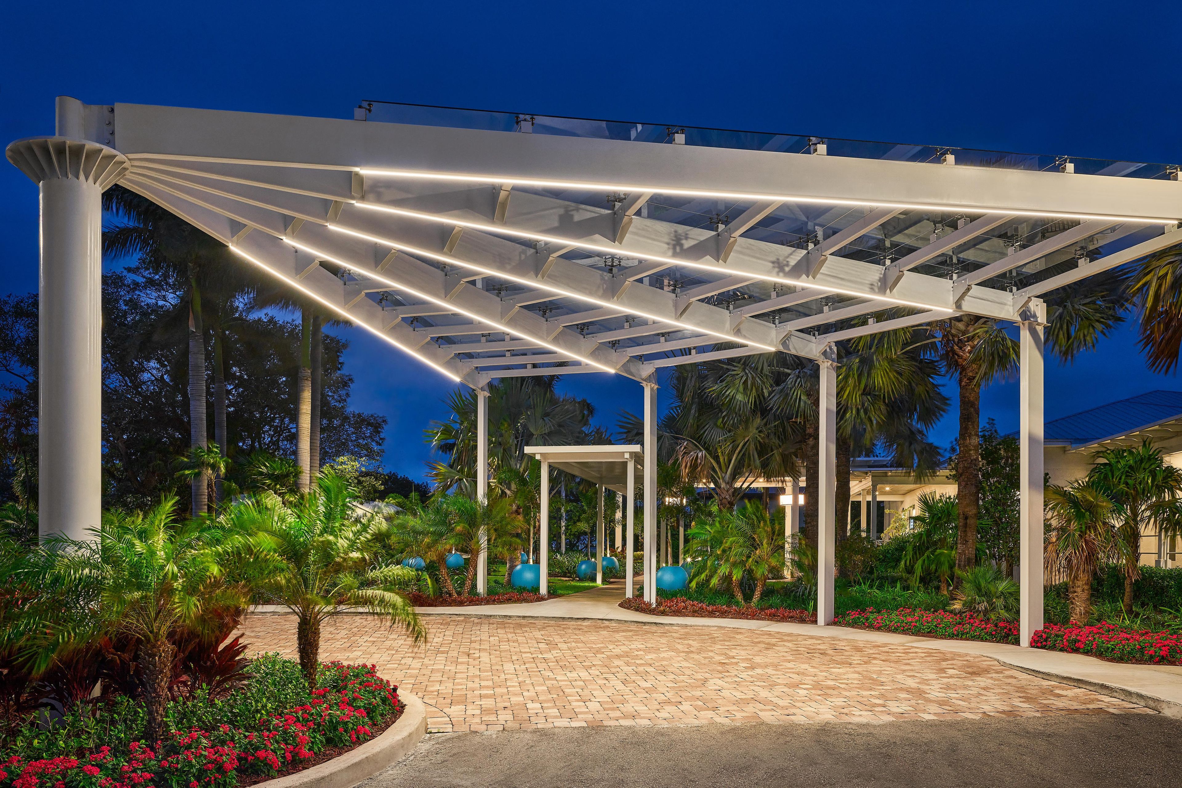 A covered outdoor area with a paved walkway, palm trees, and a building in the background.