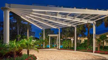 A covered outdoor area with a paved walkway, palm trees, and a building in the background.