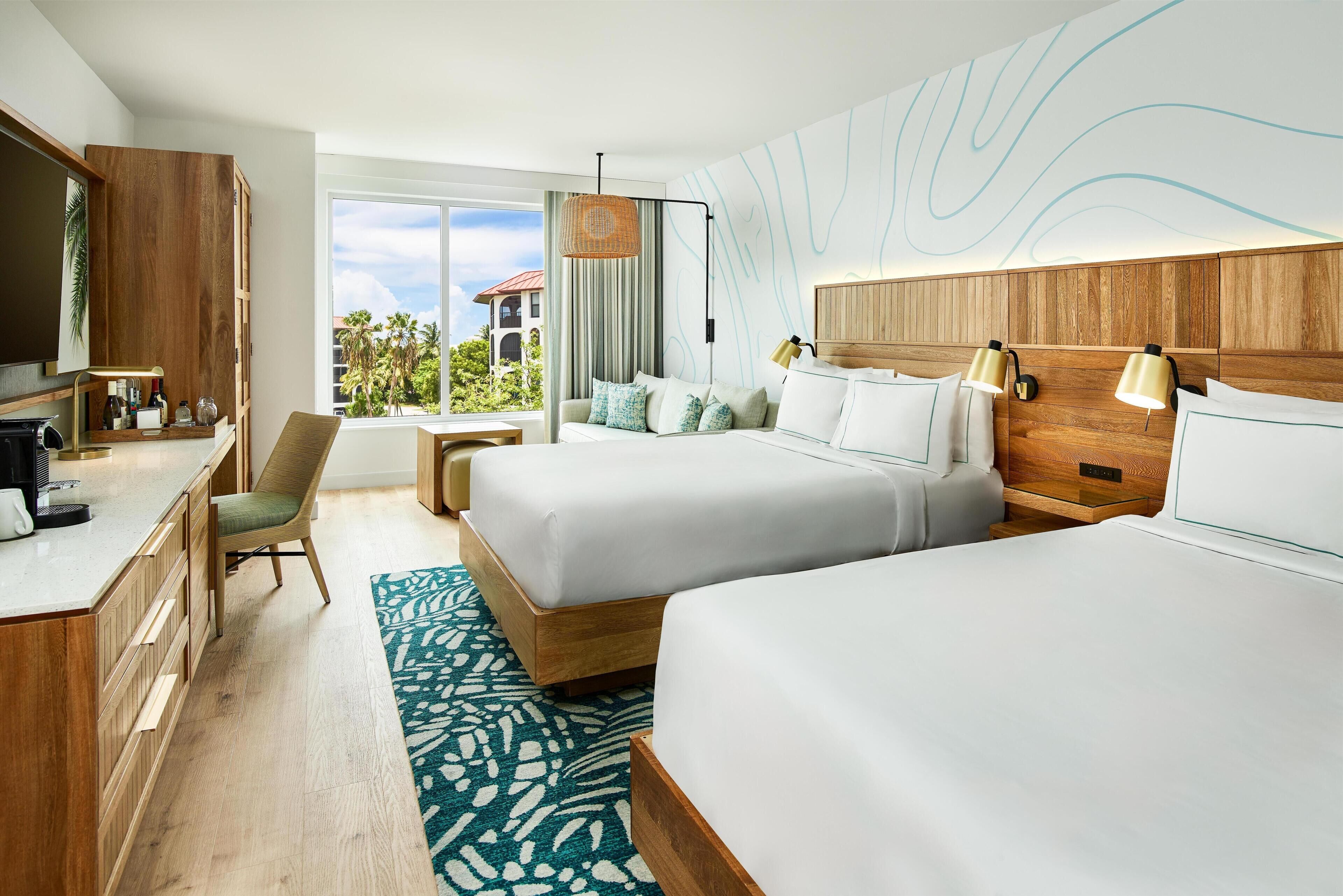 Une chambre d’hôtel avec deux lits, un bureau, une chaise, une grande fenêtre et une vue sur des palmiers.