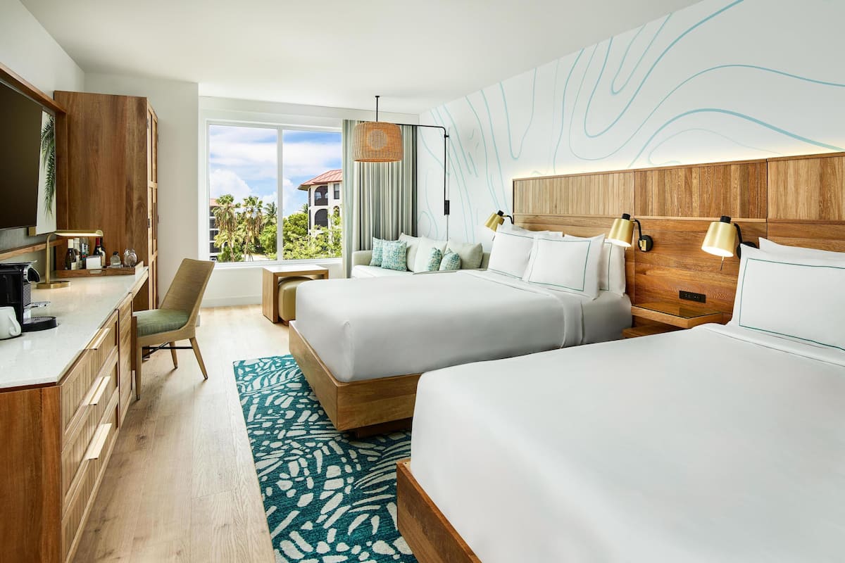 A hotel room with two beds, a desk, a chair, a large window, and a view of palm trees.