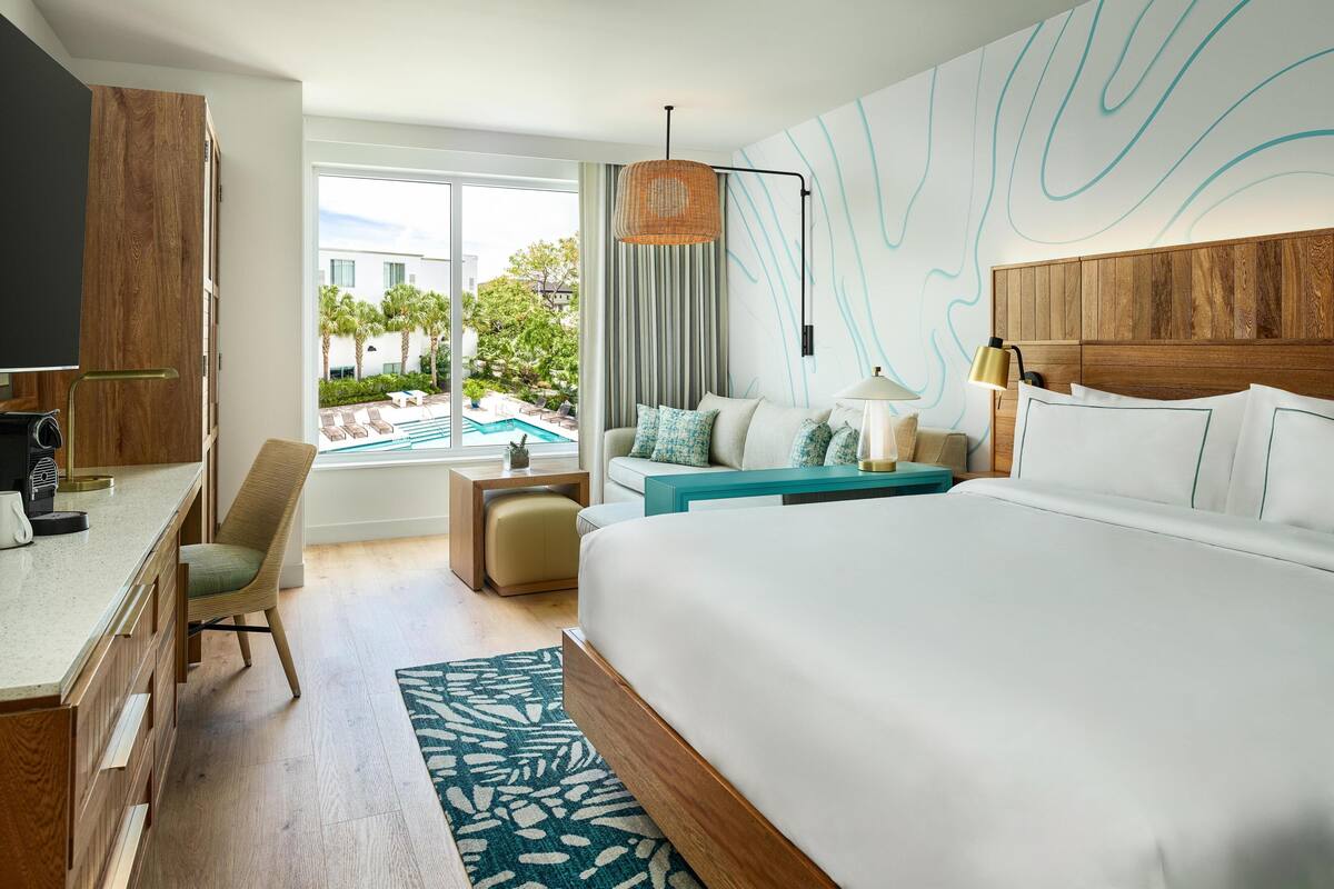 A hotel room with a large bed, a desk, a chair, a small table, and a view of an outdoor swimming pool.