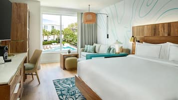 A hotel room with a large bed, a desk, a chair, a small table, and a view of an outdoor swimming pool.