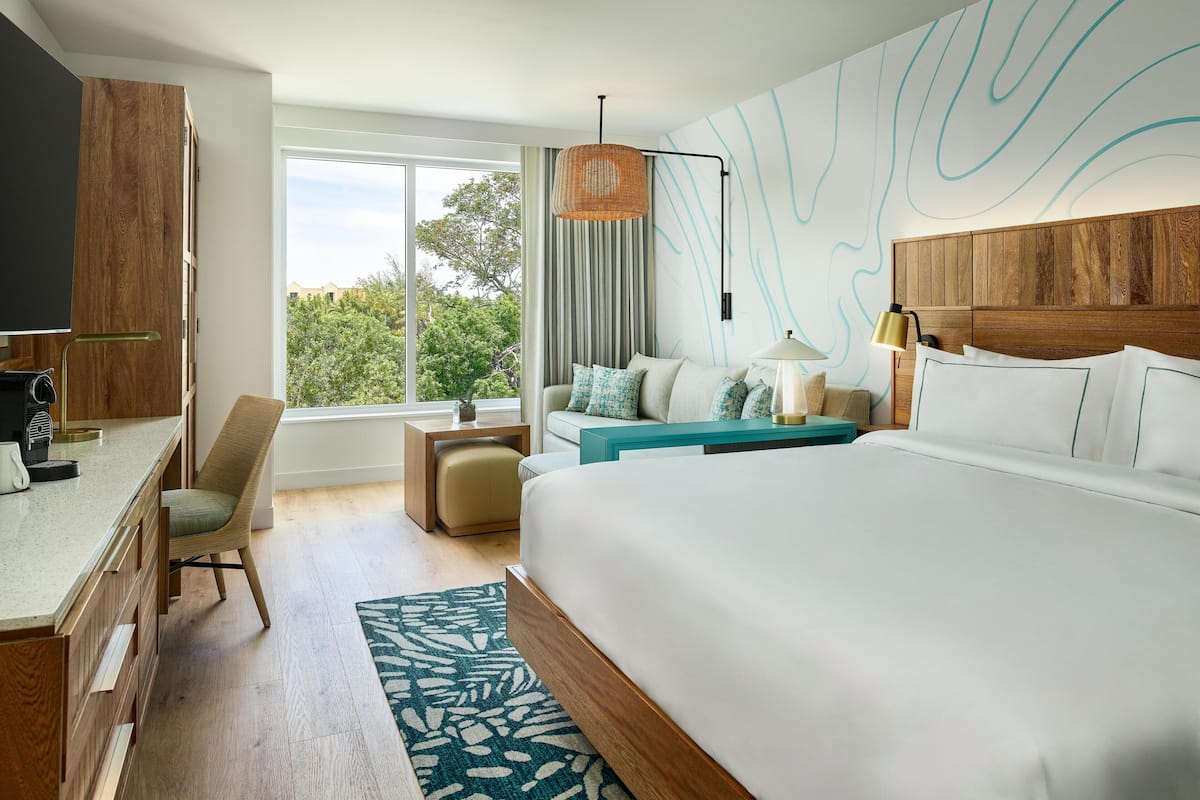 A modern hotel room with a large bed, a desk, a chair, a small table, and a view of greenery outside.