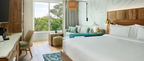 A modern hotel room with a large bed, a desk, a chair, a small table, and a view of greenery outside.