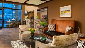 A spacious living area with a leather sofa, a coffee table, and a fireplace. There is a stone wall and a large window offering a view of the outdoors. - Meadowbrook Inn (Blowing Rock)