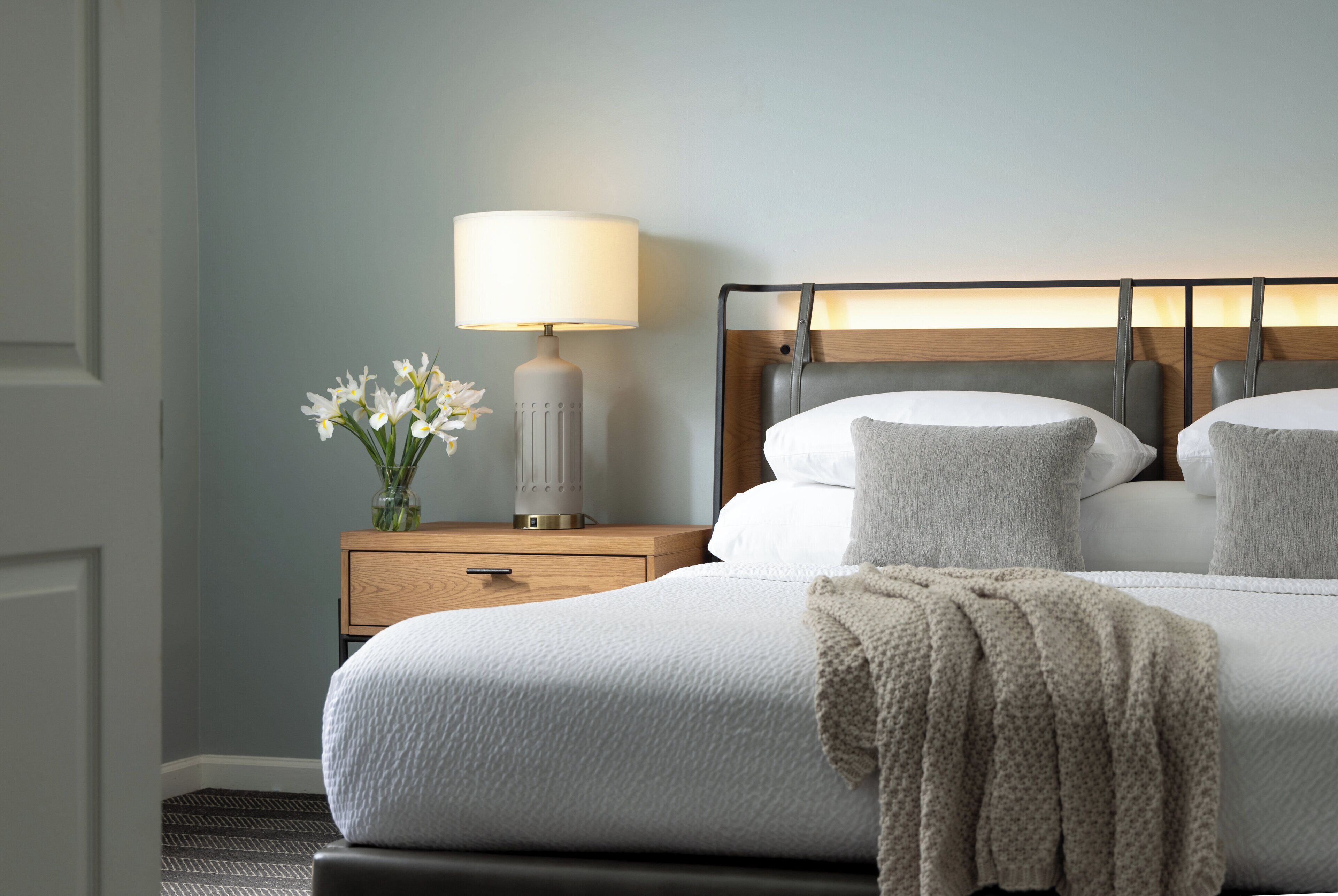 A neatly made bed with a white comforter and pillows, a bedside lamp, and a vase with white flowers on a wooden nightstand.