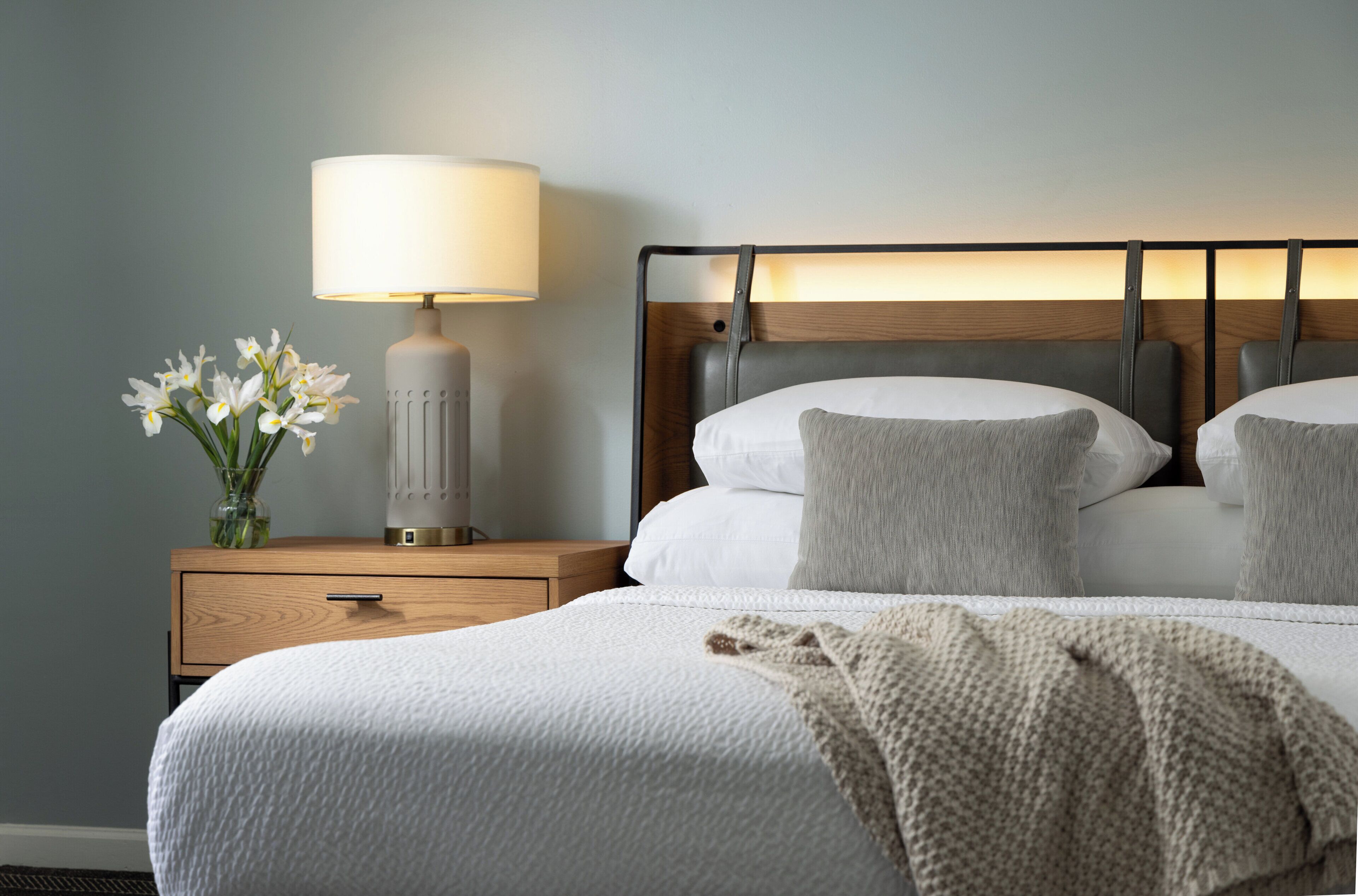 Ein ordentlich bezogenes Bett mit weißer Decke und Kissen, eine Nachttischlampe und ein Blumenvasen auf einem Holznachttisch.