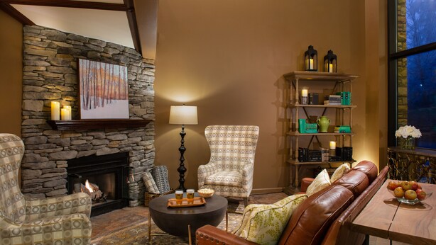 A cozy living room with a stone fireplace, leather sofa, patterned armchair, and a coffee table with drinks.