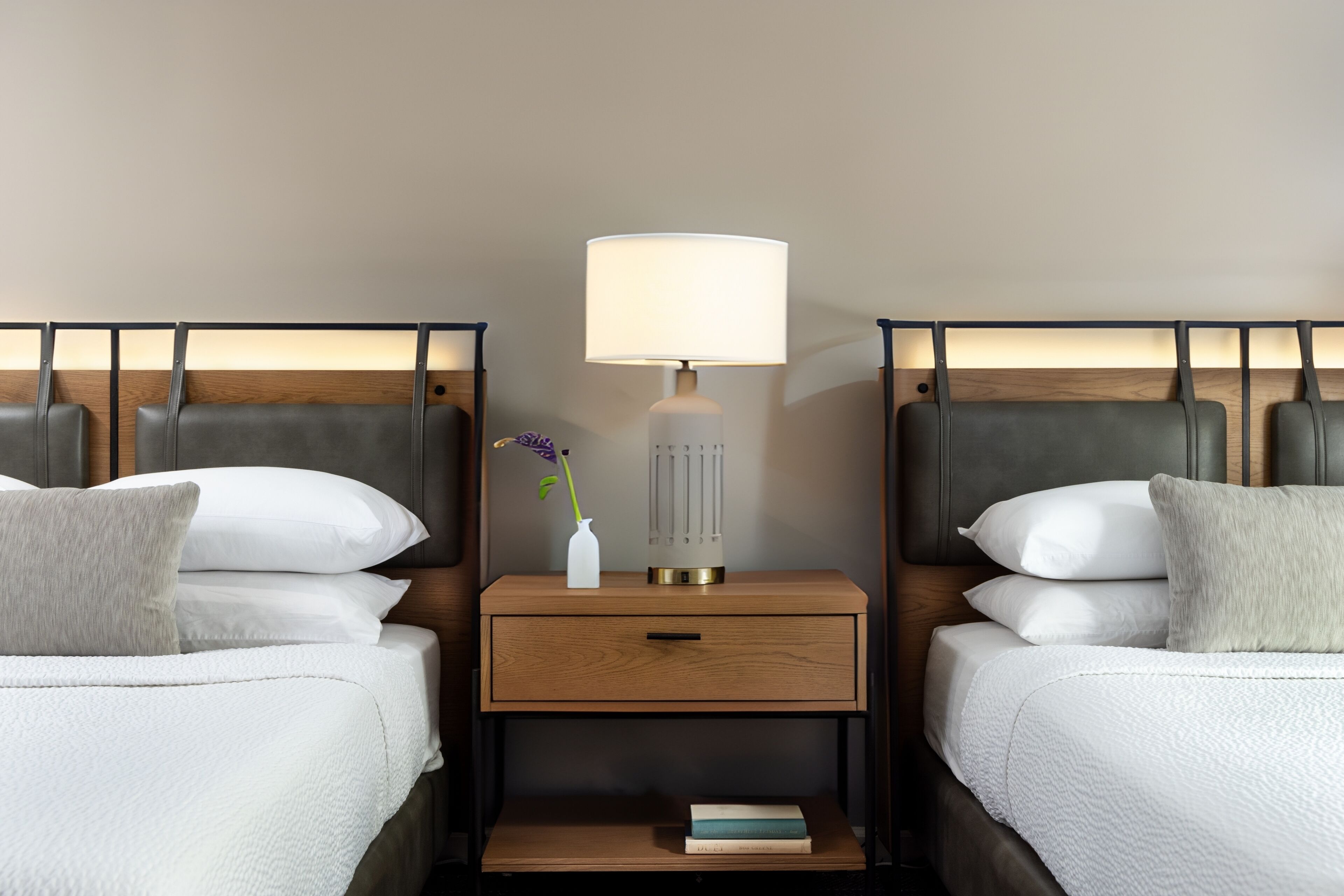 A hotel room with two beds, a nightstand with a lamp, a vase with a flower, and a book on the nightstand.