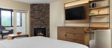 A bedroom with a large bed, a stone fireplace, a TV mounted on the wall, and a desk with a chair.