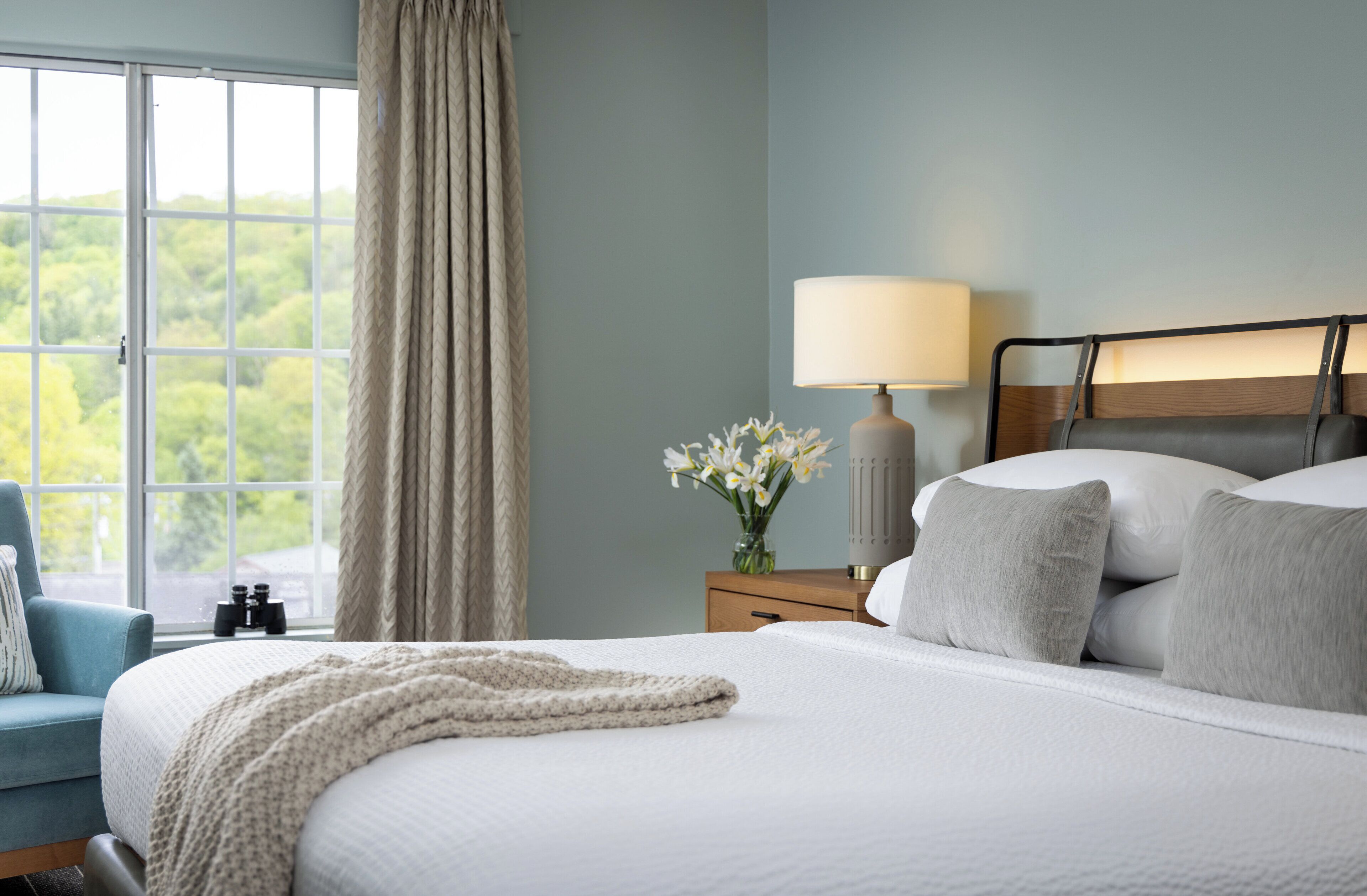 A bedroom with a large bed, a nightstand with a lamp, a vase with flowers, and a view of greenery through the window.