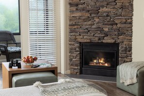 A cozy living room with a stone fireplace, a comfortable seating area, and a table with a bowl of fruit and a remote control.