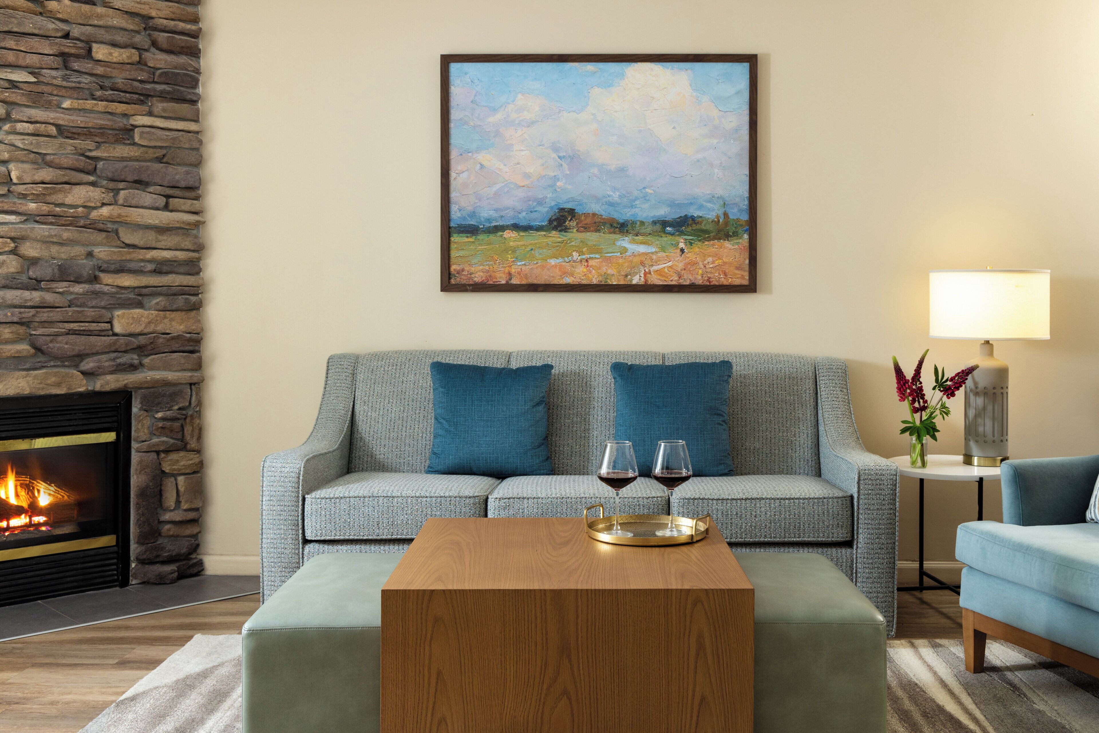 Een woonkamer met een stenen openhaard, een grijze bank met blauwe kussens, een houten salontafel en een schilderij aan de muur.